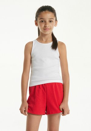 Jeune fille aux cheveux foncés attachés en queue de cheval, portant un débardeur blanc et un short rouge, debout devant un fond blanc.
