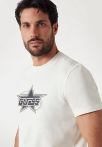 Fehér pamut póló, amelyen egy nagy szürke csillag grafika található, a "GUESS" felirattal vastag betűtípusban, kerek nyakkivágással és rövid ujjakkal.