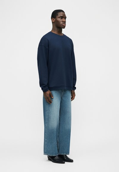 Νέος άντρας που στέκεται, φοράει σκούρο μπλε φαρδύ φούτερ, φαρδιά ανοιχτό μπλε τζιν και μαύρα μποτάκια με μυτερή μύτη σε απλό φόντο.