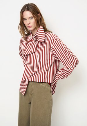 Vrouw draagt een rood-wit verticaal gestreepte blouse met een grote strik bij de kraag en olijfgroene broek, staand met één hand op de heup.
