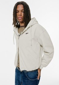 Beige zip-up hættetrøje med snorhætte, frontlommer og et broderet logo, båret over en brunt og creme-stribet skjorte.