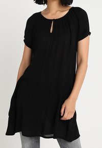 Tunique noire à manches courtes, avec un col rond orné d'une découpe en forme de goutte et un tissu subtilement texturé. Coupe ample.