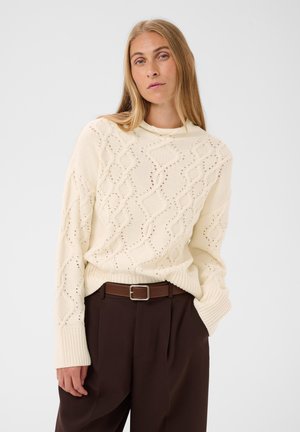 Blond kvinde iført en cremefarvet strikket sweater med diamantmønster og brune bukser med høj talje og bælte, poserende mod en hvid baggrund.