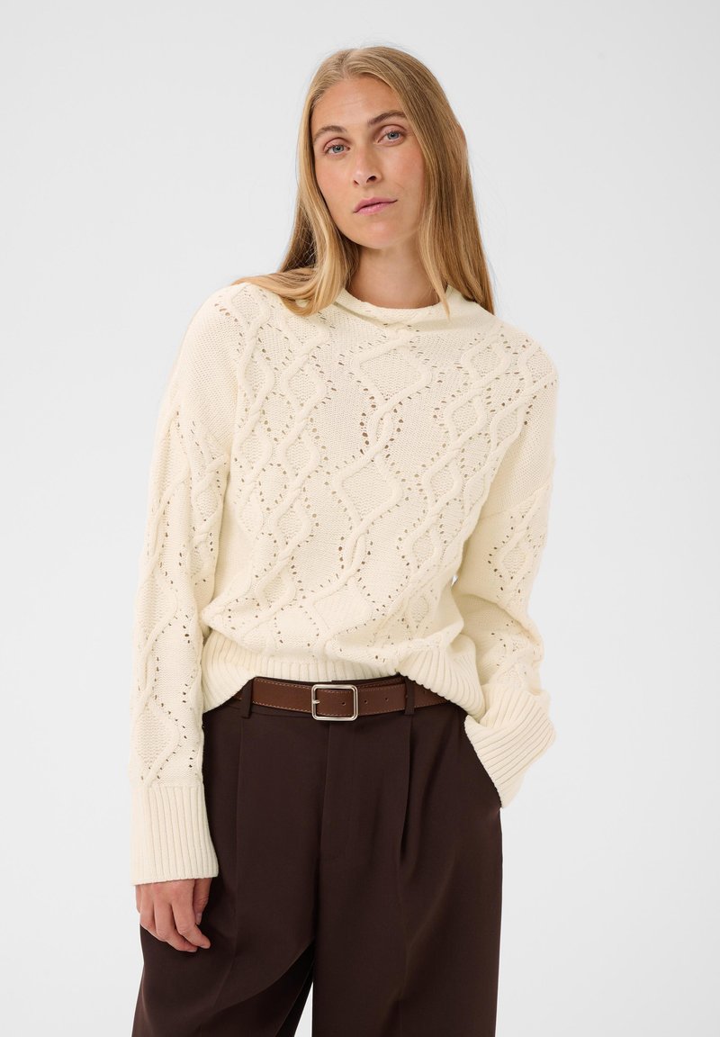 Femme blonde portant un pull en maille crème avec un motif losange et un pantalon marron taille haute avec ceinture, posant devant un fond blanc.
