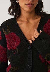 Zwarte bloemen cardigan gemaakt van zacht breimateriaal. Beschikt over grote rode rozenpatronen, een diepe V-hals en zwarte knopen met een rozenontwerp.