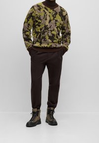 Tikkupullot, jossa on masksipainatus vihreän, ruskean ja beigen sävyissä, yhdistettynä tummanruskeisiin slim fit -haalareihin ja ruskeisiin nauhakenkiin.