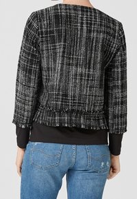 Musta-valkoinen tweed-takki, jossa on laatikkomainen muoto, tekstuuri kankaassa ja 3/4-hihat. Ominaisuuksiin kuuluu helma, joka kerääntyy vyötärölle.