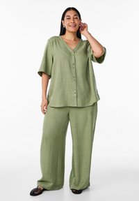 Completo in due pezzi di un verde chiaro e strutturato, composto da una camicia a maniche corte con bottoni e pantaloni larghi, entrambi con una vestibilità comoda. Sandali neri completano il look.