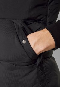 Main insérée dans la poche avant d'une veste noire matelassée avec un détail de bouton-pression sur le rabat de la poche.