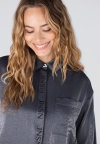 Glanzend donkergrijs shirt van gladde stof, met een kraag, een borstzak en een verticale logodetail langs de knopenlijst.