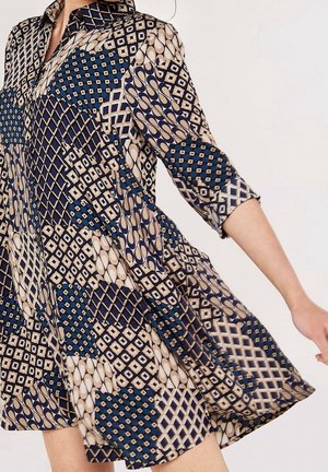 Vrouw die een losvallende jurk draagt met gemengde geometrische patronen in marineblauw, beige en wit, met driekwart mouwen.
