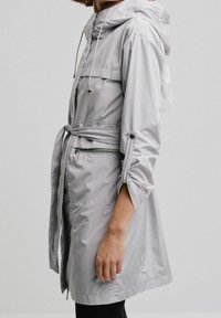 Veste imperméable gris clair avec capuche, poches zippées, taille cintrée et manches retroussées. Texture lisse avec un design élégant et moderne.