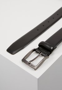 Svart lærbelte med en jevn finish, rektangulær metallspenne og flere hull for justering. Spennen har merket preget.