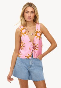 Top corto rosa con un profundo escote en V y patrones de sol en naranja, con un lazo delantero y tejido suave. Combinado con pantalones cortos de mezclilla.