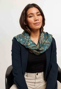 Écharpe paisley turquoise en tissu doux, drapée autour du cou, associée à un blazer sombre et une chemise noire. Pantalons beige clair.