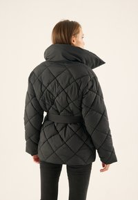 Black quilted jacket with a high collar, belted waist, and diagonal diamond pattern. Soft material, medium length, and relaxed fit.