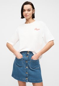 Νεαρή γυναίκα με κοντά σκούρα μαλλιά, φοράει λευκό T-shirt με το κείμενο "Amour" και μπλε τζιν φούστα με κουμπιά μπροστά και μεγάλες τσέπες.