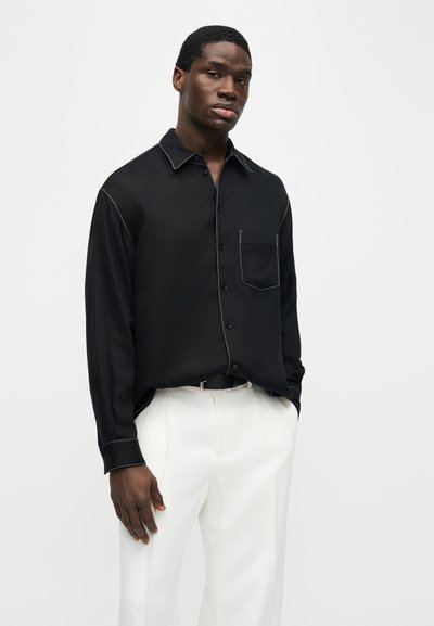 Νεαρός άνδρας που φοράει μαύρο πουκάμισο με κουμπιά και λευκές ραφές, καθώς και λευκό παντελόνι, στέκεται μπροστά σε ένα απλό φωτεινό φόντο.