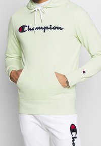 Světle zelená mikina s kapucí, přední kapsou, bílými tkaničkami a vyšitým logem "Champion" v černé a červené barvě. Obsahuje detaily s žebrovaným manžetami.