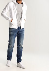 Sudadera blanca con cremallera sobre una camiseta gris, combinada con jeans azules y zapatillas blancas. La sudadera tiene un bolsillo frontal y cordones ajustables.