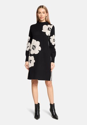 Femme portant une robe noire mi-longue ornée de grands motifs floraux blancs, à manches longues, col montant, et des bottines noires.