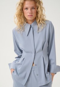 Camicia azzurra chiara a maniche lunghe con un grande collo e chiusura con bottoni. Realizzata in tessuto morbido con una vestibilità rilassata e dettaglio della tasca.