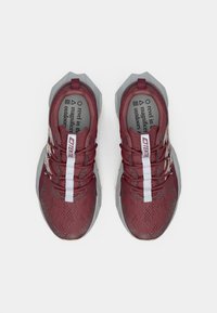 Un paio di scarpe sportive color bordeaux con suole grigie angolari, strisce bianche per i lacci verticali e soletta stampata su uno sfondo bianco uniforme.