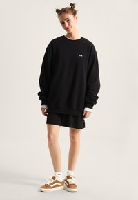 Black oversized sweatshirt with white logo, layered over a black skirt. Model wears brown and cream sneakers with white socks.