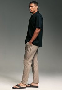 Camicetta nera a maniche corte con colletto, pantaloni slim-fit beige chiaro, infradito nere. Modello in posizione laterale su uno sfondo neutro.