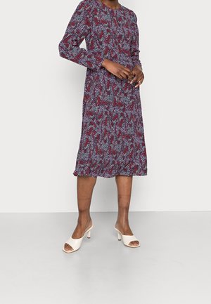 Vrouw draagt een lange mouw, knie-lange jurk met een abstract patroon in rood, wit en blauw, gecombineerd met witte open-toe sandalen met hak.