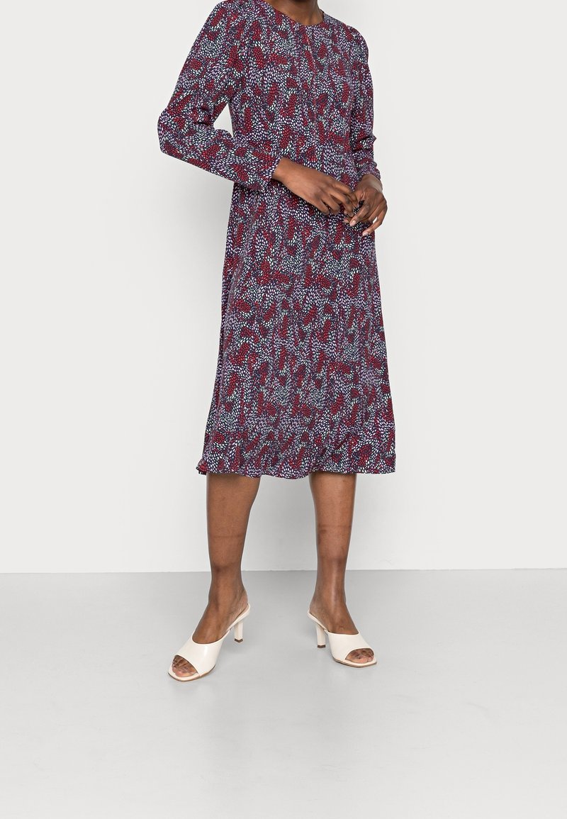 Femme portant une robe à manches longues, longueur genou, avec un motif abstrait rouge, blanc et bleu, associée à des sandales blanches ouvertes à talons.