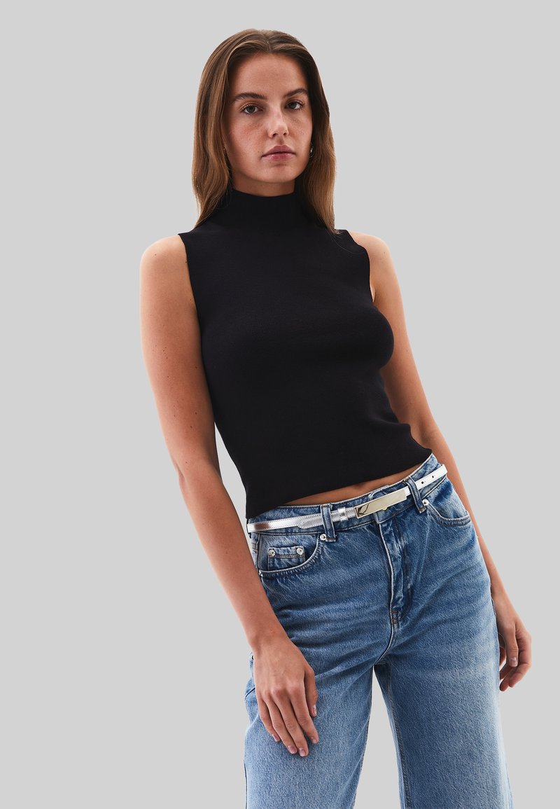 Top negro sin mangas de canalé con un cuello alto, combinado con jeans de mezclilla azul claro y un cinturón con acentos plateados. Fondo gris.
