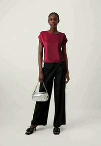 Žena koja nosi crvenu bluzu s kratkim rukavima, crne široke hlače, crne sandale s potpeticom i drži malu srebrnu torbicu.