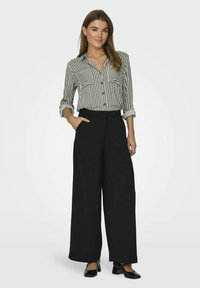 Gestreepte overhemd in zwart en wit, gecombineerd met wijde zwarte broek. De outfit heeft opgerolde mouwen en zijzakken.