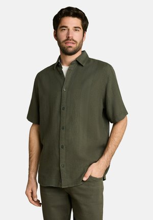 Man met baard die een donkergroen overhemd met korte mouwen en knopen en bijpassende broek draagt, poseert met één hand in zijn zak tegen een witte achtergrond.