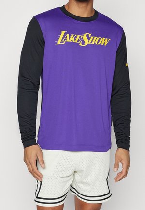 Mężczyzna ubrany w fioletową sportową koszulkę bez rękawów z żółtym napisem „LakeShow” na czarnej koszulce z długim rękawem i białe szorty do koszykówki.