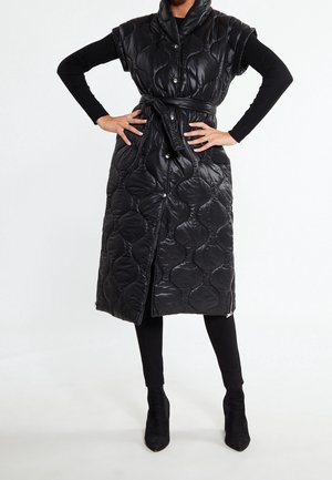 Personne portant une longue veste noire matelassée sans manches avec ceinture par-dessus un haut noir à manches longues, des leggings noirs et des bottines noires, les mains sur les hanches.