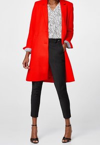 Röd skräddarsydd kappa över en vit mönstrad blus, kombinerad med svarta figurnära byxor och svarta sandaler, med en stilren och modern design.