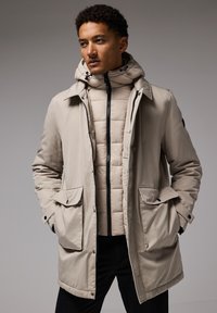 Chaqueta parka beige con un alto cuello, dos bolsillos frontales y una capa interior acolchada extraíble. Se pueden ver sus telas texturizadas y cremalleras negras.