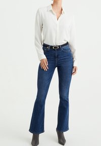 Weißes Langarmhemd mit strukturierten Punkten, kombiniert mit hoch geschnittenen dunkelblauen Jeans. Das Outfit wird mit einem schwarzen Gürtel mit einer silbernen Schnalle ergänzt.