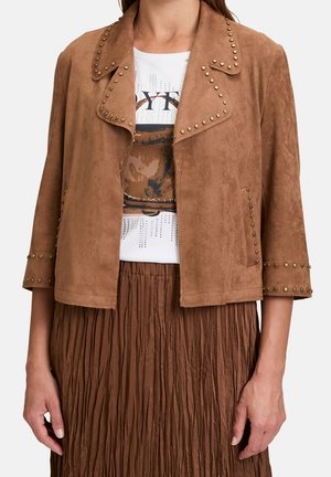 Braune Wildleder-Cropped-Jacke mit Nietenbesatz über einem weißen T-Shirt mit Grafik und einem plissierten braunen Rock.