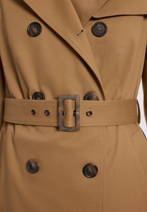 Płaszcz trencz koloru wielbłądziego, dwurzędowy, z dużymi ciemnymi guzikami i pasem w tym samym kolorze z prostokątną klamrą w wzór bursztynu.