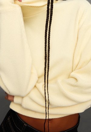 Persona vistiendo un suéter crema acanalado con largas trenzas negras colgando por delante, manos escondidas bajo las mangas.