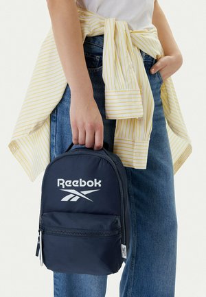 Persona vestida con vaqueros azules y camiseta blanca con una camisa a rayas amarilla atada alrededor de la cintura, sosteniendo una pequeña mochila Reebok azul marino.