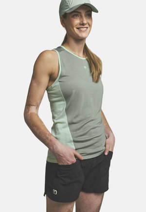 Lächelnde Frau mit grauem ärmellosem Oberteil, schwarzen Shorts und hellgrauer Kappe, steht mit den Händen in den Taschen vor weißem Hintergrund.