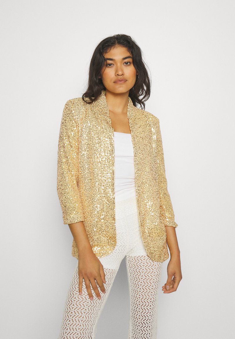 Blazer in paillettes dorées con un taglio morbido, apertura frontale e maniche a tre quarti, abbinato a pantaloni bianchi strutturati.