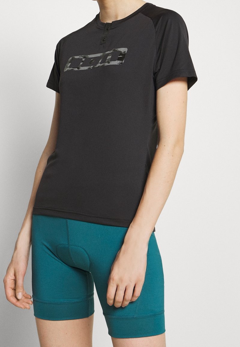 Femme portant un haut de sport noir à manches courtes avec un logo et un short de cyclisme rembourré bleu sarcelle, debout devant un fond blanc.