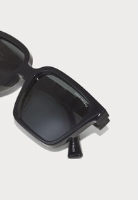 Ochelari de soare negri, supradimensionați, cu lentile dreptunghiulare, rame din plastic groase și detalii cu logo pe braço. Design elegant, minimalist.