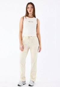 Top senza maniche color crema con bordi marroni e logo, abbinato a pantaloni cargo beige con tasche frontali. Il modello indossa sneakers grigie.