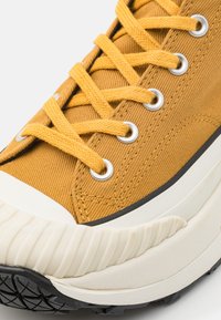 Yellow canvas sneaker featuring round silver eyelets and textured laces, with a white rubber toe cap and a black rubber treaded sole.